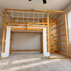 May include: A wooden loft bed frame featuring a white rope safety net along the top bunk, a vertical climbing wall with multi-colored handholds, and a side ladder. The structure includes white support panels and a slatted base for a mattress.