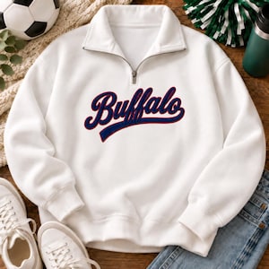 May include: A white long-sleeve quarter-zip pullover sweatshirt featuring the word Buffalo embroidered in a navy blue script font with a red outline across the chest. The garment has a high collar, a silver metal zipper, and ribbed cuffs.