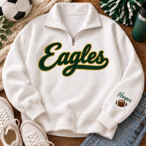 May include: A white quarter-zip pullover sweatshirt featuring the word Eagles in large, dark green cursive script with a yellow outline across the chest. The left sleeve displays the name Harper in green text above a small brown football graphic.