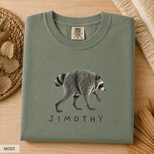 May include: A moss green crew neck t-shirt featuring a detailed black and white illustration of a raccoon walking. Below the raccoon, the name JIMOTHY is printed in simple, dark capital letters. The shirt has a Comfort Colors brand label.