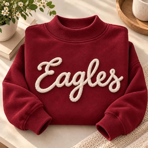 May include: A deep red long-sleeve sweatshirt featuring a mock neck collar and ribbed cuffs. The word Eagles is written across the center in a raised, textured, cream-colored script font that appears to be made of thick yarn or chenille.