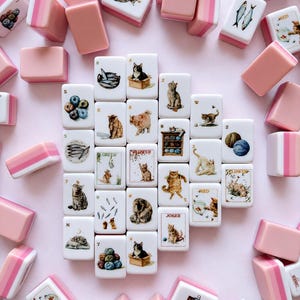 May include: A collection of rectangular mahjong tiles featuring illustrations of cats, fish, yarn, and flowers. The tiles have white faces with colorful graphics and pink-toned backs. Some tiles display text including Joker, Red, Green, and Flower.