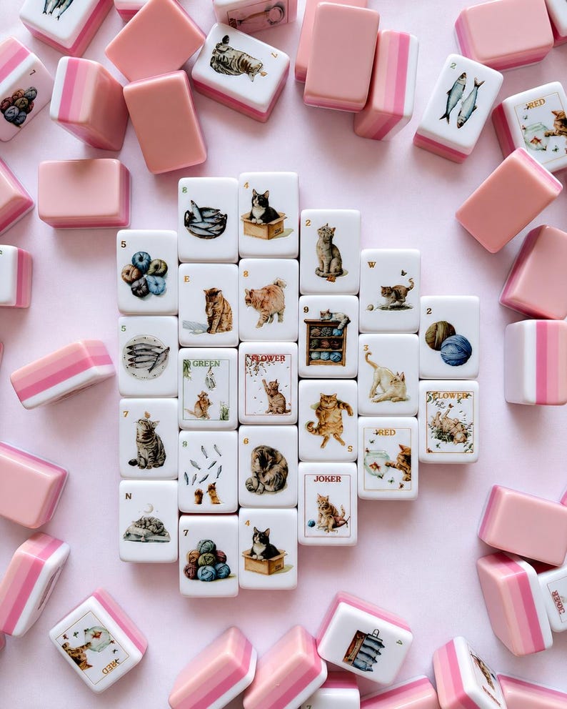 May include: A collection of rectangular mahjong tiles featuring illustrations of cats, fish, yarn, and flowers. The tiles have white faces with colorful graphics and pink-toned backs. Some tiles display text including Joker, Red, Green, and Flower.