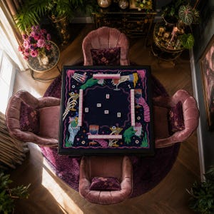 May include: A square mahjong table featuring a dark blue felt surface with a colorful graphic print of skeletal hands, eyeballs, and cocktail glasses. The table includes four rows of light pink mahjong tiles and several scattered tiles.