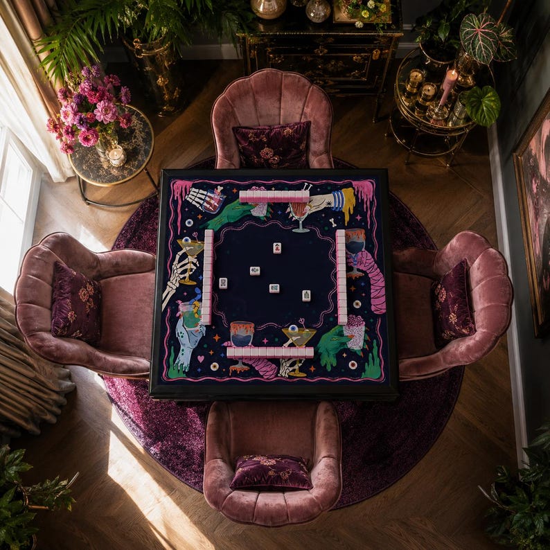 May include: A square mahjong table featuring a dark blue felt surface with a colorful graphic print of skeletal hands, eyeballs, and cocktail glasses. The table includes four rows of light pink mahjong tiles and several scattered tiles.
