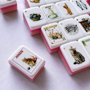 May include: A collection of white Mahjong tiles with pink and white layered bases. Each tile features detailed illustrations of cats, fish, yarn, and soap. One tile is labeled JOKER, another SOAP, and another FLOWER. The tiles are arranged in a scattered pattern.