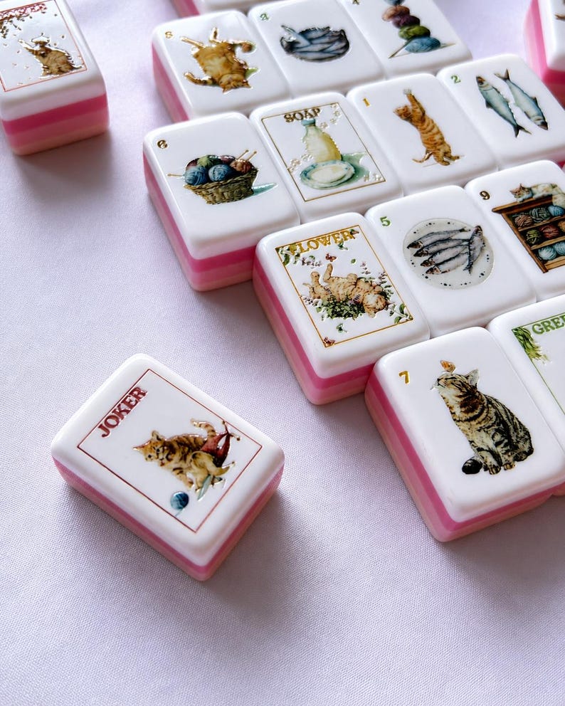 May include: A collection of white Mahjong tiles with pink and white layered bases. Each tile features detailed illustrations of cats, fish, yarn, and soap. One tile is labeled JOKER, another SOAP, and another FLOWER. The tiles are arranged in a scattered pattern.