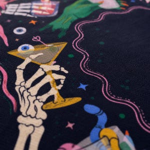 May include: A black fabric featuring a colorful graphic print of a skeleton hand holding a martini glass with an eyeball inside. The design includes additional elements like a green hand, pink wavy lines, blue stars, and small colorful dots.