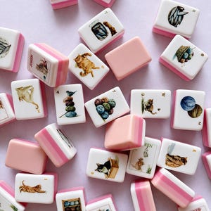 May include: A collection of rectangular mahjong tiles featuring white faces with colorful illustrations of cats, yarn balls, and fish, paired with pink and white striped backs. Some tiles display text including Joker, Soap, Green, and letters W and E.