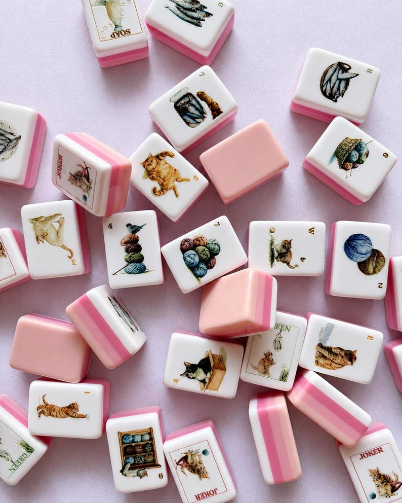 May include: A collection of rectangular mahjong tiles featuring white faces with colorful illustrations of cats, yarn balls, and fish, paired with pink and white striped backs. Some tiles display text including Joker, Soap, Green, and letters W and E.