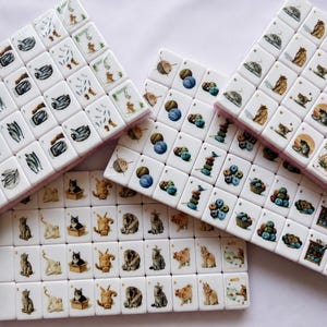 May include: A set of white rectangular mahjong tiles featuring colorful illustrations of cats, balls of yarn, and fish. Each tile displays a unique cat pose, stacked yarn, or fish pattern, with some tiles showing small numbers or letters in the corners.
