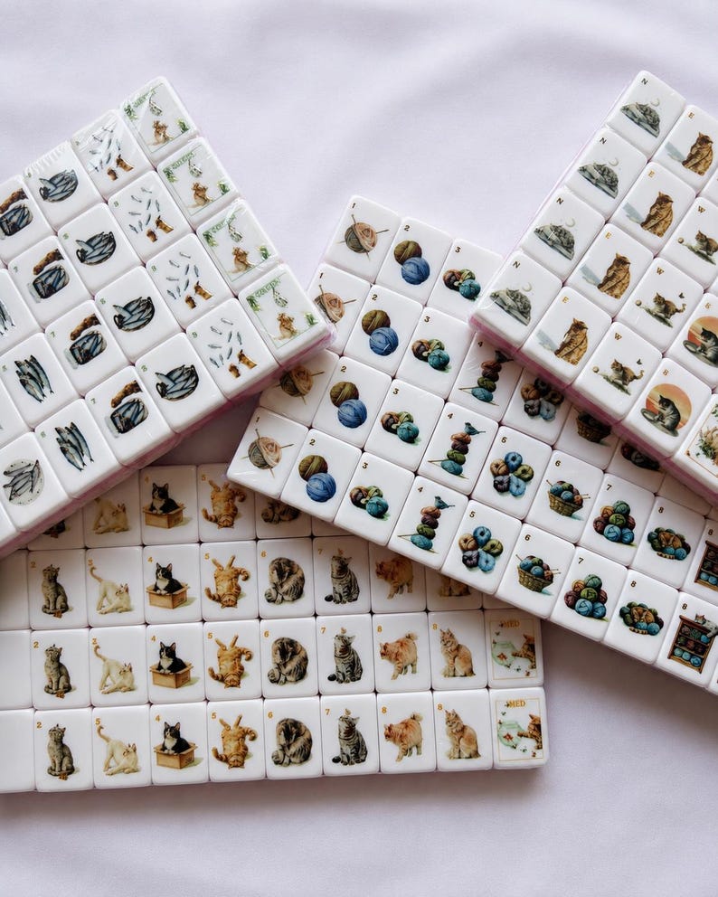 May include: A set of white rectangular mahjong tiles featuring colorful illustrations of cats, balls of yarn, and fish. Each tile displays a unique cat pose, stacked yarn, or fish pattern, with some tiles showing small numbers or letters in the corners.