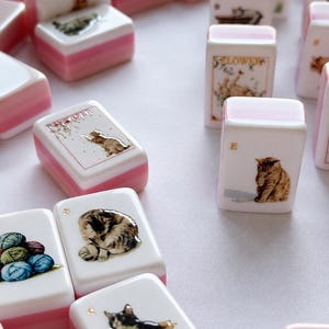 May include: A collection of rectangular mahjong tiles featuring various cat illustrations and yarn balls. Each white tile has a pink base and displays detailed imagery, including cats in boxes, cats playing, and colorful yarn, with some tiles marked with letters.
