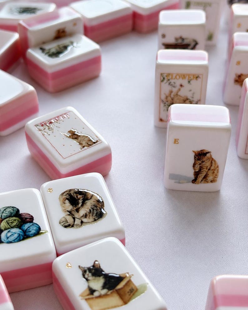 May include: A collection of rectangular mahjong tiles featuring various cat illustrations and yarn balls. Each white tile has a pink base and displays detailed imagery, including cats in boxes, cats playing, and colorful yarn, with some tiles marked with letters.