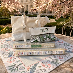 May include: A cream-colored canvas tote bag and a clear zippered pouch labeled TILES containing green and white mahjong tiles. Two long, translucent, scalloped-edge tile racks hold rows of mahjong tiles on a floral-patterned game mat.