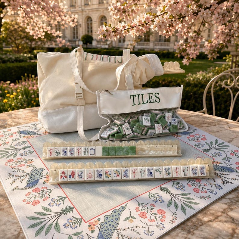 May include: A cream-colored canvas tote bag and a clear zippered pouch labeled TILES containing green and white mahjong tiles. Two long, translucent, scalloped-edge tile racks hold rows of mahjong tiles on a floral-patterned game mat.