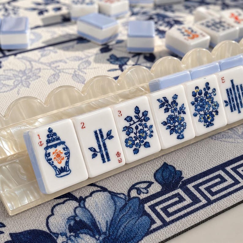 Może przedstawiać: Zestaw białych i jasnoniebieskich płytek do gry w Mahjong ułożonych na falowanej, perłowo-białej podstawce. Płytki ozdobione są niebiesko-pomarańczowymi ilustracjami kwiat&oacute;w, bambusa i wazon&oacute;w z czerwonymi cyframi na niebiesko-białej macie.