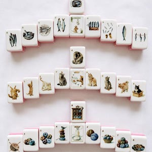 May include: A collection of white rectangular mahjong tiles arranged in three horizontal rows. The tiles feature colorful illustrations of fish, cats, and yarn balls, each marked with a small black number. Some tiles include text labels like GREEN, RED, and SOAP.
