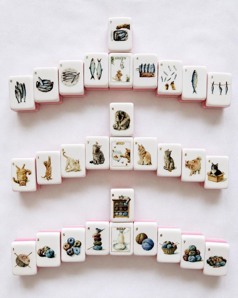 May include: A collection of white rectangular mahjong tiles arranged in three horizontal rows. The tiles feature colorful illustrations of fish, cats, and yarn balls, each marked with a small black number. Some tiles include text labels like GREEN, RED, and SOAP.