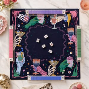 May include: A square mahjong mat featuring a dark blue center with five white tiles. The border displays colorful illustrations of skeleton hands, zombie hands, and mummy hands holding various cocktail glasses, eyeballs, and stars.
