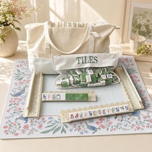 May include: A Mahjong set featuring a cream canvas tote bag, a clear zippered pouch labeled TILES containing green and white tiles, four translucent tile racks, and a rectangular play mat with a floral and bird pattern.