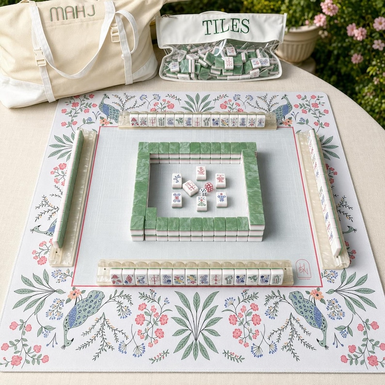 May include: A Mahjong set featuring a white play mat with a floral and peacock pattern, four tile racks, and a square wall of green and white tiles. Includes a canvas bag labeled MAHJ and a clear pouch labeled TILES with extra game pieces.