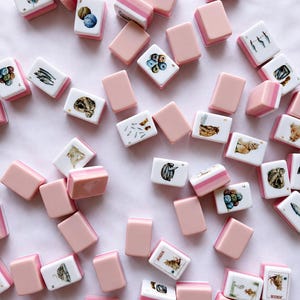 May include: A collection of scattered mahjong tiles featuring a white face with various illustrations including cats, fish, and yarn balls, paired with a solid pastel pink back. Some tiles are marked with numbers or the word Joker in black text.