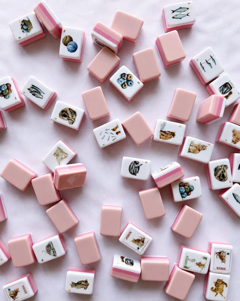 May include: A collection of scattered mahjong tiles featuring a white face with various illustrations including cats, fish, and yarn balls, paired with a solid pastel pink back. Some tiles are marked with numbers or the word Joker in black text.