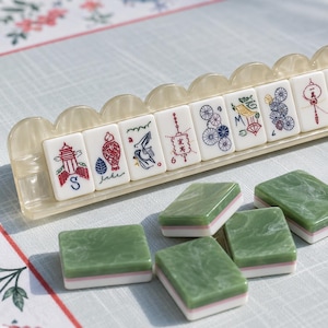 May include: A set of mahjong tiles featuring white tiles with colorful illustrations of lanterns, flowers, and symbols arranged in a scalloped beige rack, alongside several green and white marbled tiles scattered in the foreground.