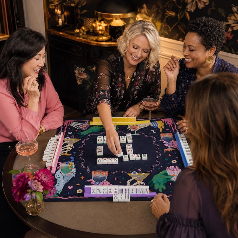 May include: A square mahjong mat featuring a dark navy blue background with colorful illustrations of skeletons, cocktails, and eyeballs. The mat includes four plastic tile racks in yellow, pink, blue, and purple, with white mahjong tiles arranged.