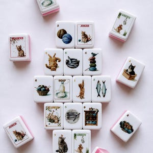 May include: A collection of rectangular mahjong tiles with white faces and pink backs. Each tile features detailed illustrations of cats, yarn balls, fish, and the words SOAP, JOKER, FLOWER, or GREEN. The tiles are arranged in a central cluster.