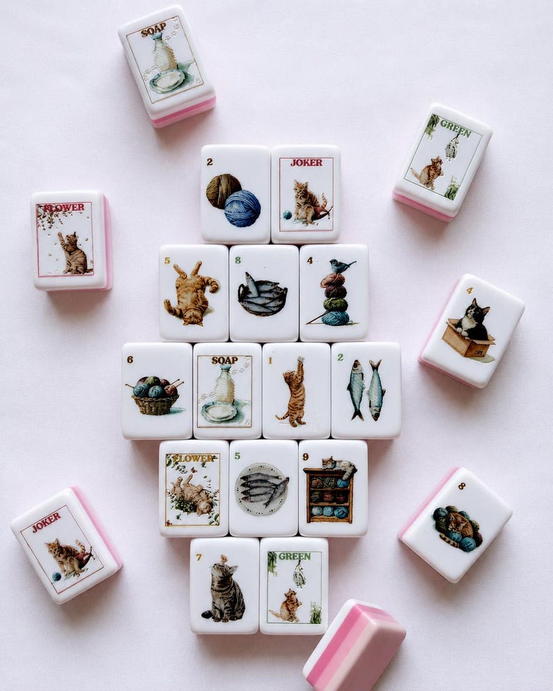 May include: A collection of rectangular mahjong tiles with white faces and pink backs. Each tile features detailed illustrations of cats, yarn balls, fish, and the words SOAP, JOKER, FLOWER, or GREEN. The tiles are arranged in a central cluster.