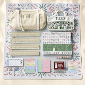 May include: A mahjong game set featuring a cream canvas bag labeled MAHJ, a clear pouch labeled TILES, four translucent scalloped racks, a set of green and white tiles, dice, score pads, and a National Mah Jongg League card on a floral mat.