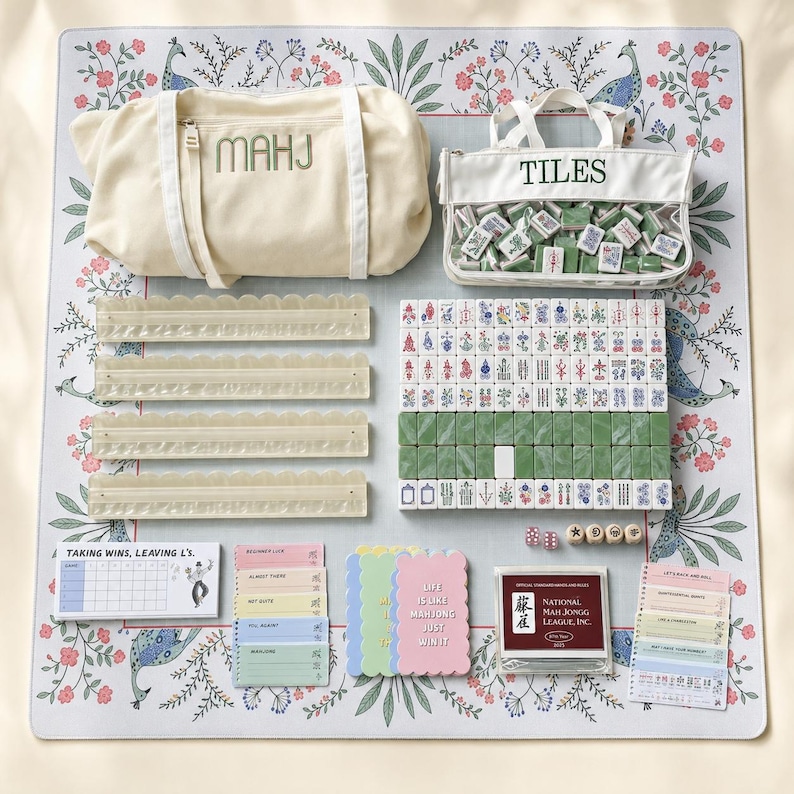 May include: A mahjong game set featuring a cream canvas bag labeled MAHJ, a clear pouch labeled TILES, four translucent scalloped racks, a set of green and white tiles, dice, score pads, and a National Mah Jongg League card on a floral mat.