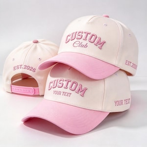 Könnte beinhalten: Zwei cremefarbene und rosafarbene Baseballkappen mit Snapback-Verschluss. Die Vorderseiten zeigen den gestickten Text CUSTOM CLUB und CUSTOM YOUR TEXT. An den Seiten befinden sich die Stickereien EST. 2026 und YOUR TEXT in Rosa.