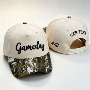 May include: Two off-white baseball caps with a camouflage pattern on the brim. The front cap features the word Gameday in black script. The back cap displays the text YOUR TEXT in black block letters and the number #40 on the side panel.