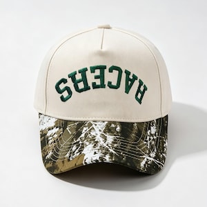 Peut inclure: Une casquette de baseball couleur crème avec le mot RACERS brodé en lettres capitales vert foncé dans un design arqué sur les panneaux avant. La visière incurvée présente un motif camouflage détaillé en vert, marron et blanc.
