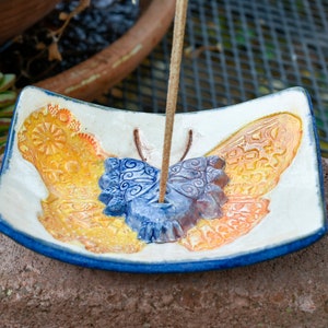 May include: A ceramic incense holder shaped like a butterfly with yellow, orange, and blue details. The holder has a hole in the center for an incense stick.