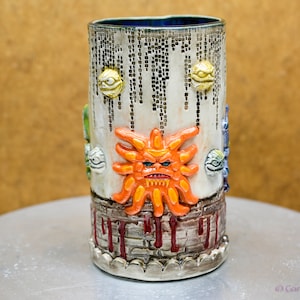 May include: A ceramic mug with a white background and a colorful design. The design features a red, orange, and yellow sun-like creature with a menacing expression. The mug also has a blue and green design with a pattern of small squares.