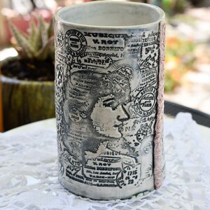 May include: A white ceramic mug with a black and white design of vintage stamps and text. The mug has a red stripe on the side. The text on the mug includes "MUSIQUE", "V. ROY", "Edition DONNEFO", "Tumelot", "1829", "ANTO", "479", "MUSIQUE", "V. ROY", "Zden BORREPOND", "PARIS-X", "1715", "23", "AU GENTRAL", "MUSIQUE", "PARIS".