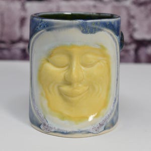 May include: A ceramic mug with a blue and white glaze. The mug has a face sculpted on the front, with closed eyes and a smiling mouth. The face is a light yellow color.