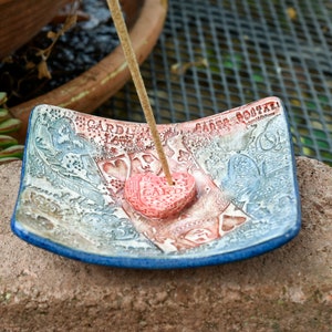 May include: A square ceramic incense holder with a blue rim and a white, red, and blue design. The holder features a heart-shaped indentation in the center and the text "GARDEN" and "LOVE" on the side. An incense stick is placed in the heart-shaped indentation.