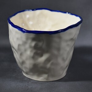 May include: White ceramic cup with a blue rim. The cup has a textured, uneven surface.