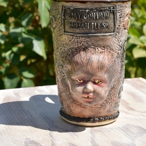 May include: A ceramic mug with a textured surface and a grotesque face sculpted on the front. The mug has a black rim and a black band around the bottom. The mug has a sign that reads "May Contain Human Flesh".
