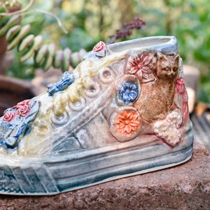 May include: A ceramic planter shaped like a sneaker with a cat and flowers sculpted on the side. The planter is painted in shades of blue, green, yellow, red, and brown.