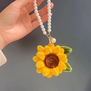 May include: A sunflower charm featuring a textured yellow petal design with a dark brown center and green leaves, attached to a white faux pearl beaded strap with a gold-toned metal clasp and a single dangling pearl accent.