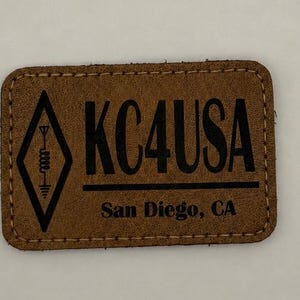 May include: A rectangular brown leather patch with rounded corners and stitched edges. The patch features a black printed design including a diamond-shaped antenna icon, the text KC4USA, and the location text San Diego, CA below a line.