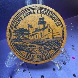May include: A circular tan leather patch featuring a black laser-engraved illustration of the Point Loma Lighthouse in San Diego. The design includes a lighthouse, a flag, a tree, and rocky cliffs, with the text Point Loma Lighthouse San Diego.