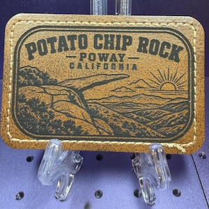 May include: A rectangular tan leather patch with rounded corners and white stitching around the border. The patch features a black laser-engraved illustration of Potato Chip Rock in Poway, California, with a sunrise and text reading POTATO CHIP ROCK.
