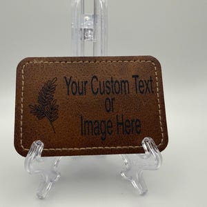 May include: A rectangular brown leather patch with rounded corners and tan stitching along the edges. The patch features a black engraved fern graphic on the left side and the black text Your Custom Text or Image Here on the right side.